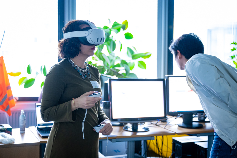 A woman is wearing VR glasses and a person next to her is looking at a computer screen.omputerbildschirm