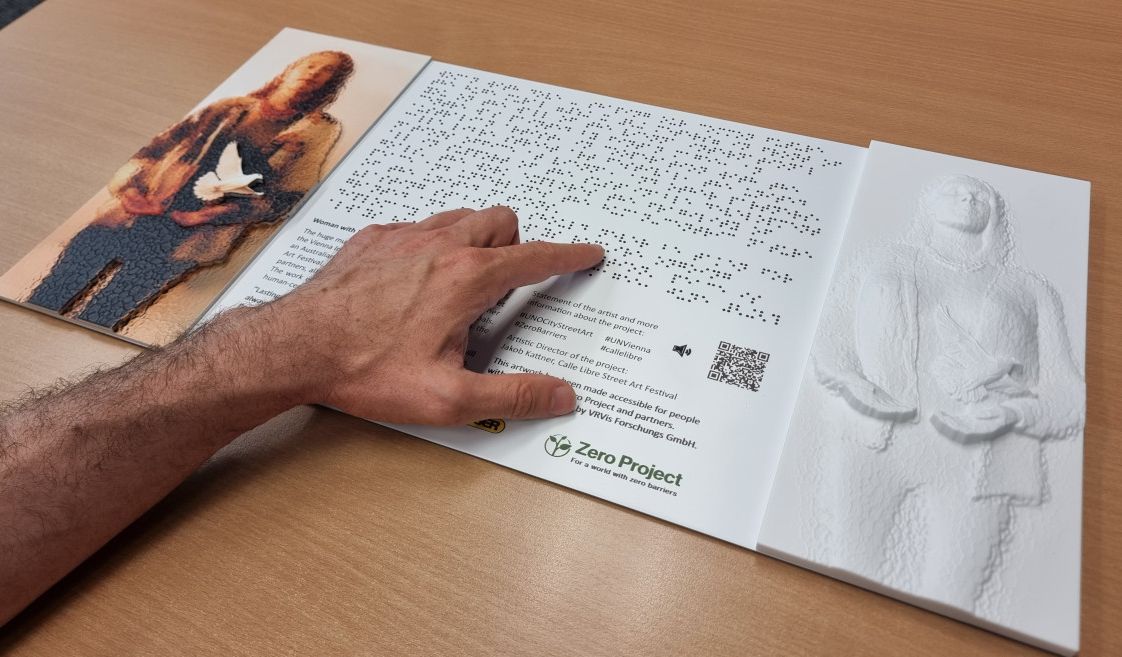 Close-up of the barrier-free information board for the mural "Woman with Dove" by Fintan Magee, a man's hand rests on the board and reads the Braille.